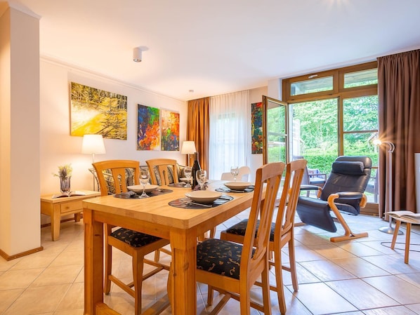 A dining area with a wooden table and chairs, a painting on the wall, and a view of greenery outside.