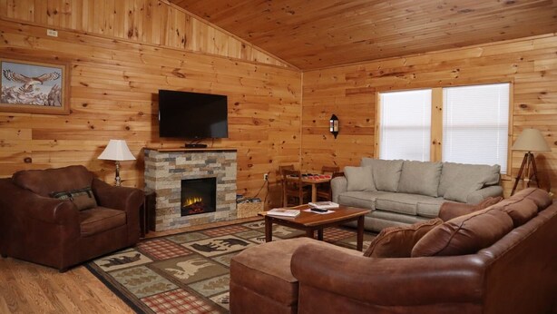 A cozy cabin interior with a stone fireplace, leather sofas, a flat-screen TV, and a dining area. - Laurel Lodge-Devil`s Bathtub/Nat Tunnel (DUFFIELD)