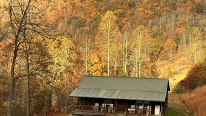 A rustic cabin with a porch and outdoor seating, surrounded by a forested hillside. - Laurel Lodge-Devil`s Bathtub/Nat Tunnel (DUFFIELD)