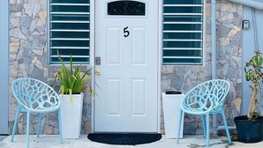 Uma porta branca com o número 5, acompanhada por duas cadeiras azuis e plantas em vasos, apoiada em uma parede de pedra.