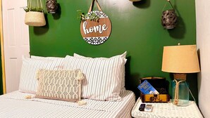 A bedroom with a green wall, striped bedding, a hanging 'home' sign, and a bedside table with a lamp and a book. - Bohemian Eco Solar Power Near Ferry To Culeb (Ceiba)
