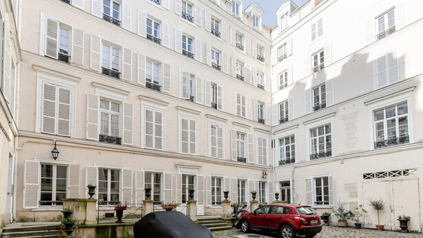 Une cour où est garée une voiture rouge devant un immeuble aux volets blancs et doté de balcons.