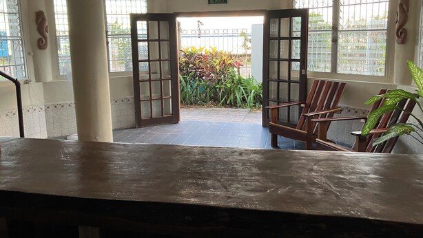 A hotel lobby with a wooden reception desk, two open doors leading outside, and a ceiling fan.