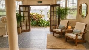 A room with two wooden chairs, a woven rug, a floor fan, and a view of a garden through an open door. - Hotel Martin  (Corozal)
