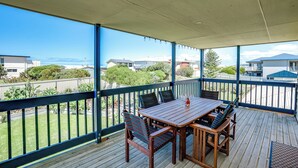 Een overdekt terras buiten met een houten tafel en stoelen, een uitzicht op de oceaan en een heldere lucht.
