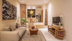 A modern living room with a sofa, coffee table, and TV stand. There is a dining area with a table and chairs, and a kitchen in the background. - Villa Kayu Pererenan (Canggu)