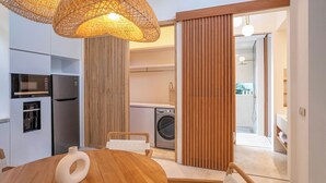A modern kitchen with a built-in oven, a refrigerator, and a washing machine. There is a wooden dining table with chairs and a hanging light fixture above. - Villa Kayu Pererenan (Canggu)