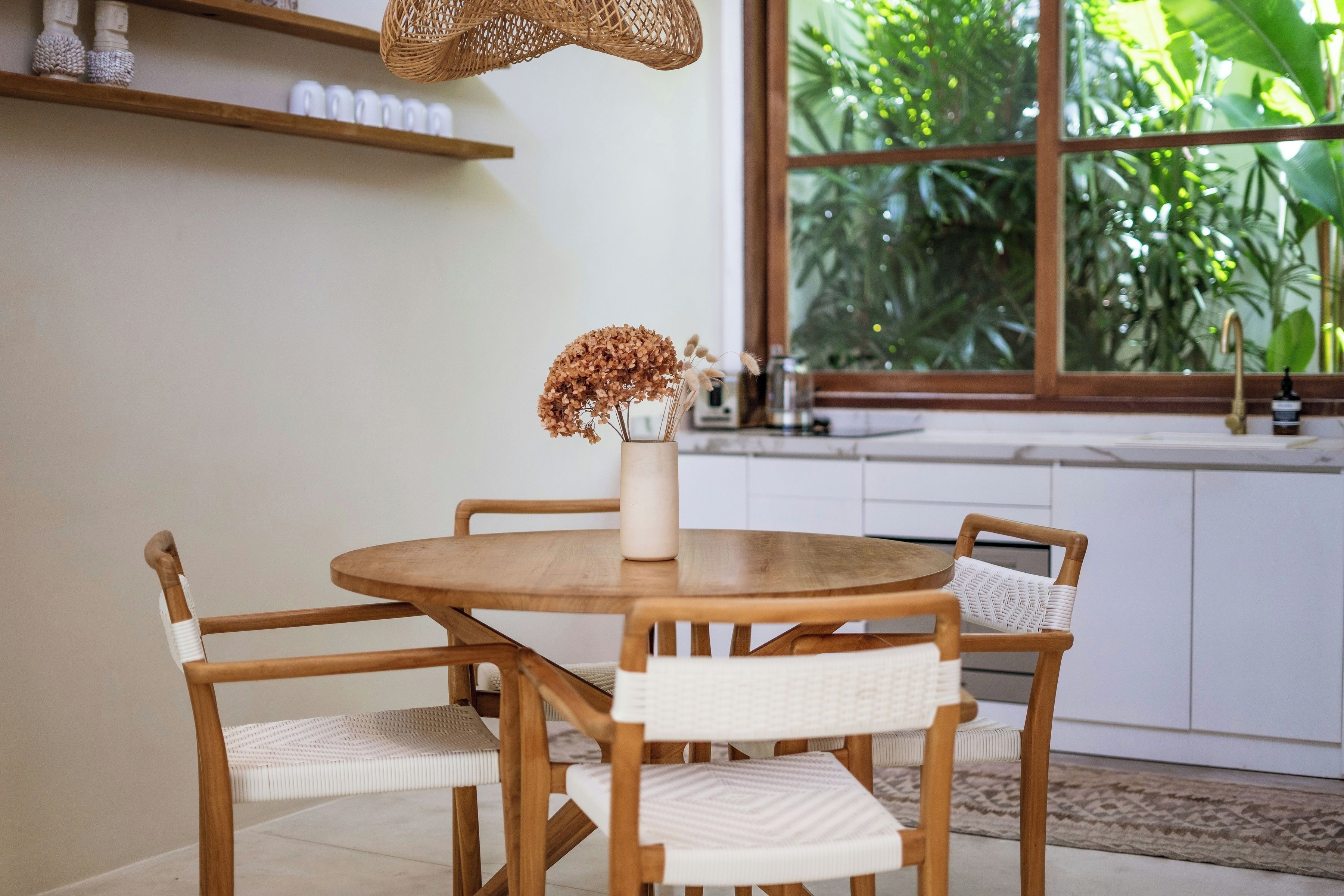 A round wooden dining table with four chairs, a vase with dried flowers, and a wicker hanging light fixture.