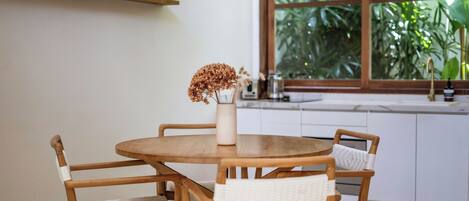 A round wooden dining table with four chairs, a vase with dried flowers, and a wicker hanging light fixture.