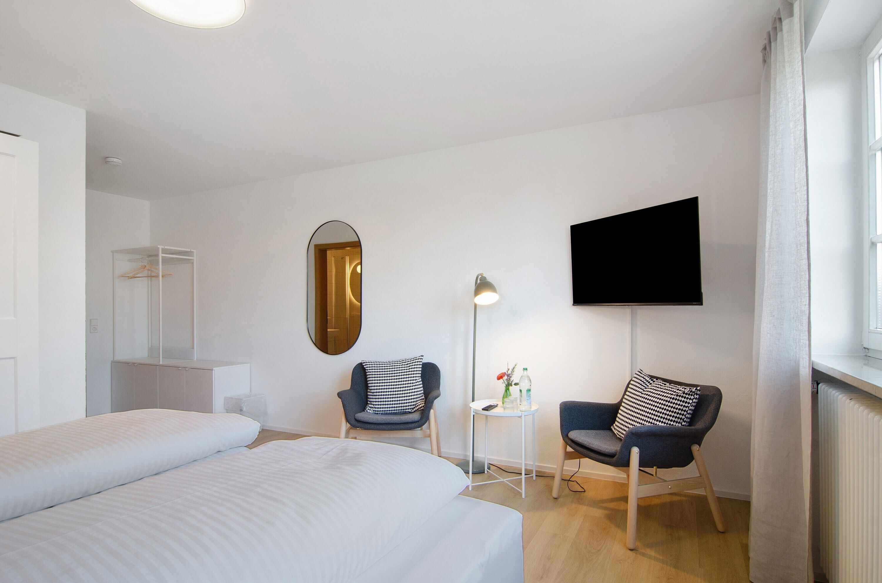 A hotel room with a bed, two chairs, a TV, a mirror, and a small table with a lamp.