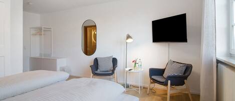 A hotel room with a bed, two chairs, a TV, a mirror, and a small table with a lamp.