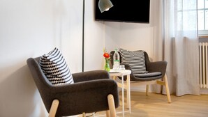 A modern living room with two armchairs, a small table with a vase and a bottle, and a floor lamp. A flat-screen TV is mounted on the wall. - Room 'Konstanz' with Shared Terrace, Shared Garden and Wi-Fi (Salem)