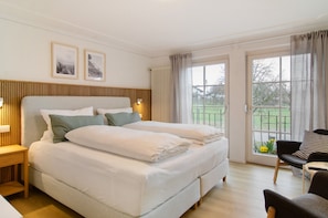 Quarto com cama grande, mesa de cabeceira, cadeira e janela com vista para o verde.