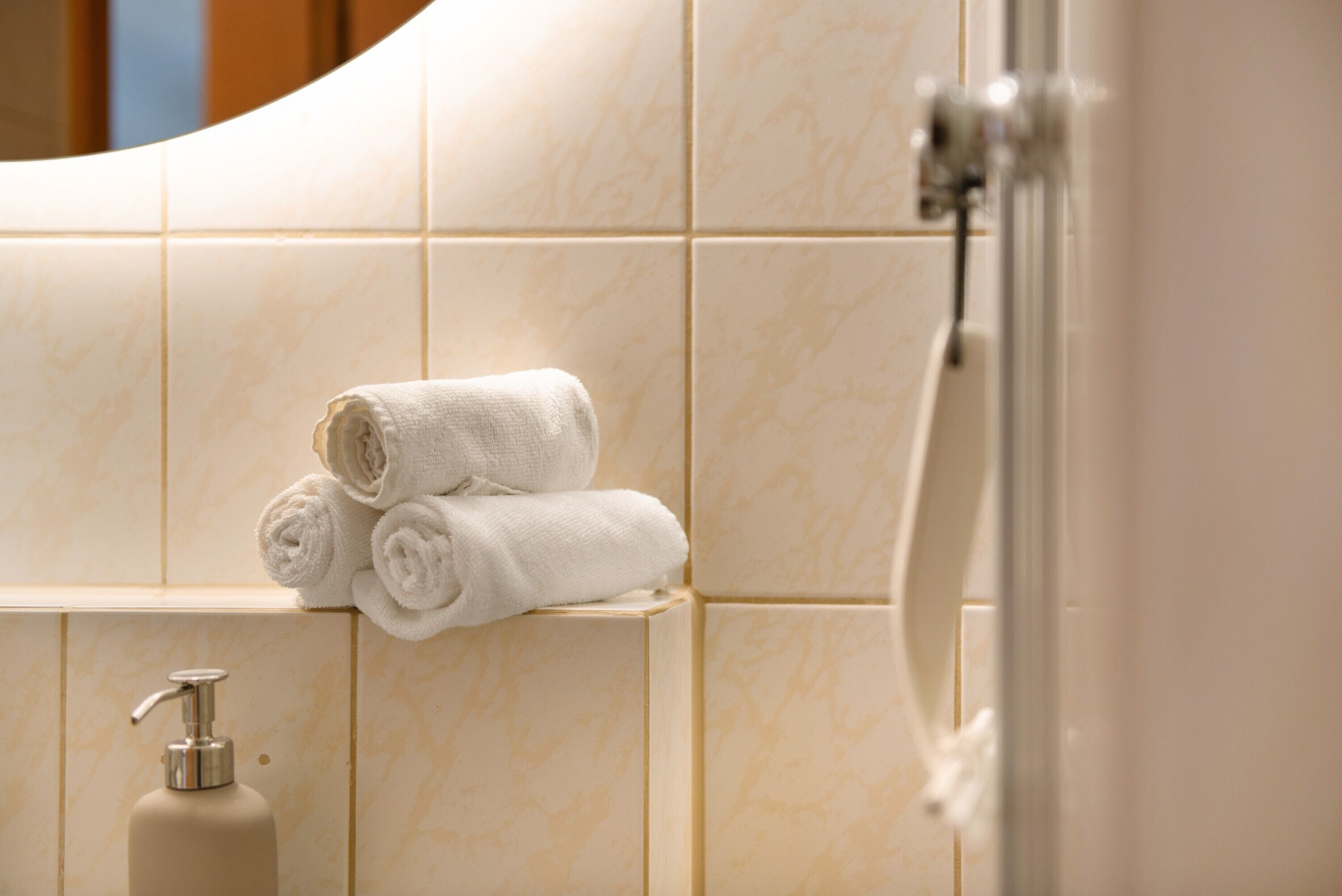 A bathroom with beige tiles, a bottle of shampoo, and rolled towels on a shelf.