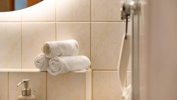 A bathroom with beige tiles, a bottle of shampoo, and rolled towels on a shelf.