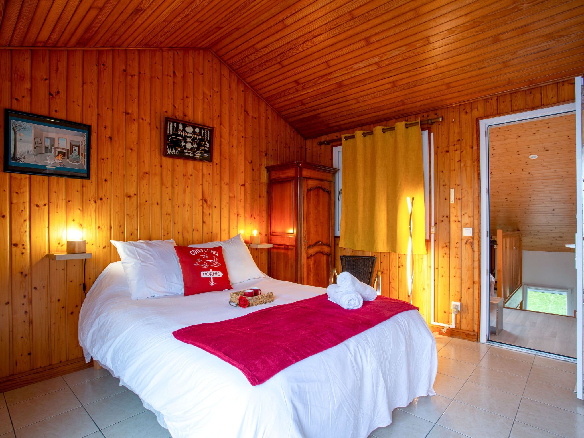 A wooden-paneled bedroom with a bed, pillows, a red throw blanket, a framed picture, and a nightstand with a lamp.