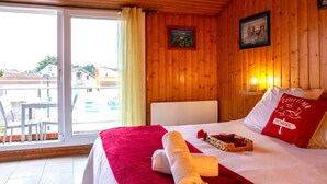 A hotel room with a wooden interior, a bed with a red blanket and pillows, a window with a view of buildings, and a table with a tray of items. - Ker Zelia by Interhome (La Bernerie-en Retz)