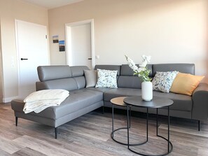 A modern living room with a grey sectional sofa, a round coffee table, and a vase of flowers. - Seaside 53° FeWo 5 - Fantastic vacation apartment with sea view in Cuxhaven for the (Cuxhaven)