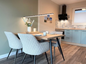 A modern kitchen with a dining area, featuring a wooden table, grey upholstered chairs, a pendant light, and a tiled backsplash.