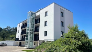 Un immeuble moderne à plusieurs étages, doté d’un puits d’ascenseur en verre, avec une voiture garée devant et une végétation luxuriante tout autour.