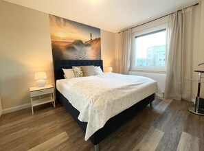 A hotel room with a large bed, a nightstand with a lamp, a window with curtains, and a cityscape view. - Seaside 53° FeWo 5 - Fantastic vacation apartment with sea view in Cuxhaven for the (Cuxhaven)