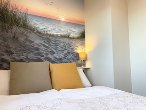 A bedroom with a bed, pillows, a nightstand, a lamp, and a wall mural of a beach scene.