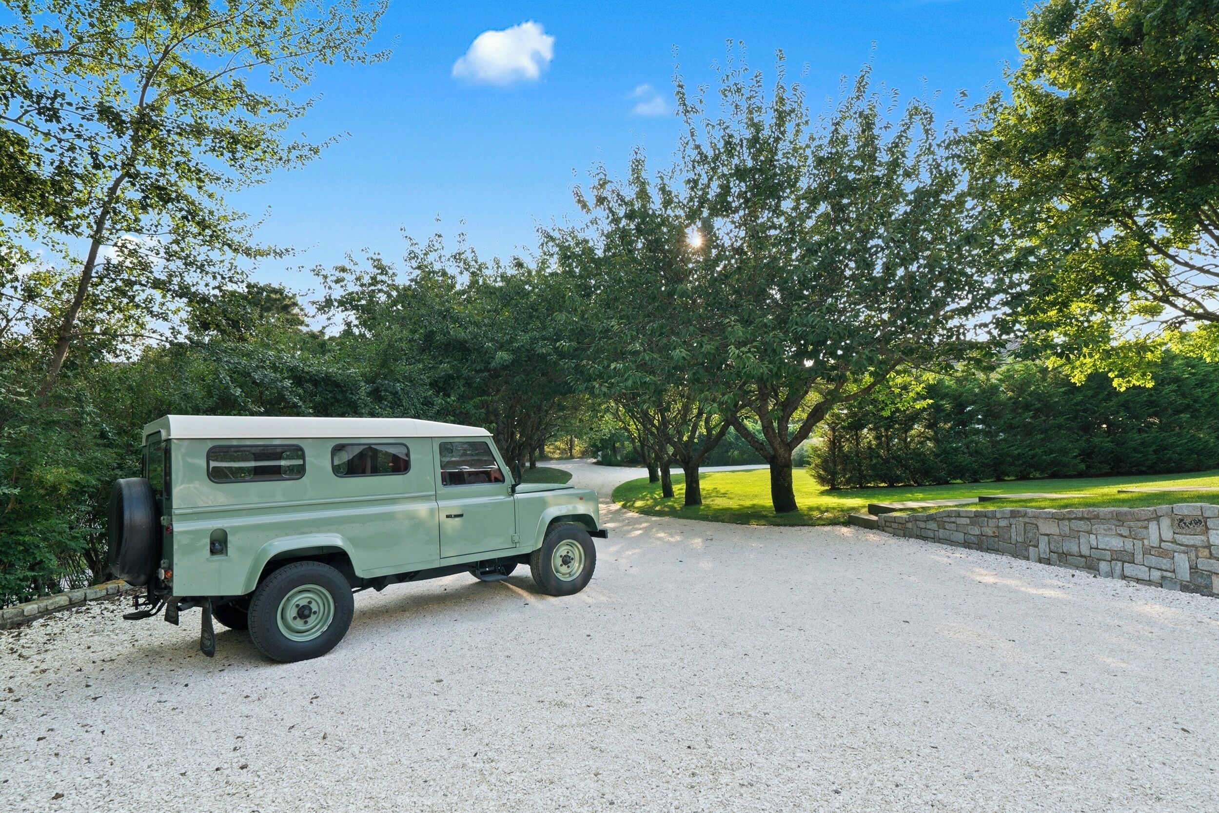 A vintage green van parked on a gravel driveway in a park-like setting with trees and a stone wall.