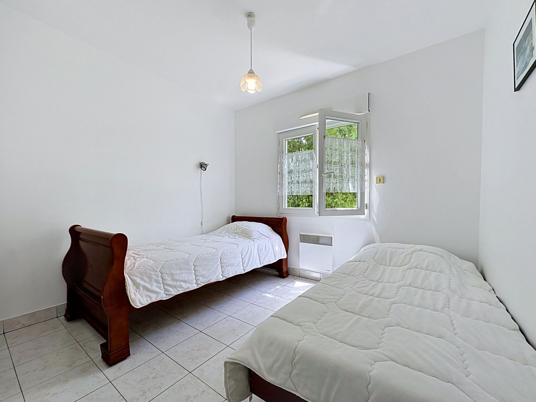 Una habitación con dos camas, una ventana con cortinas de encaje, una lámpara en el techo y un radiador.