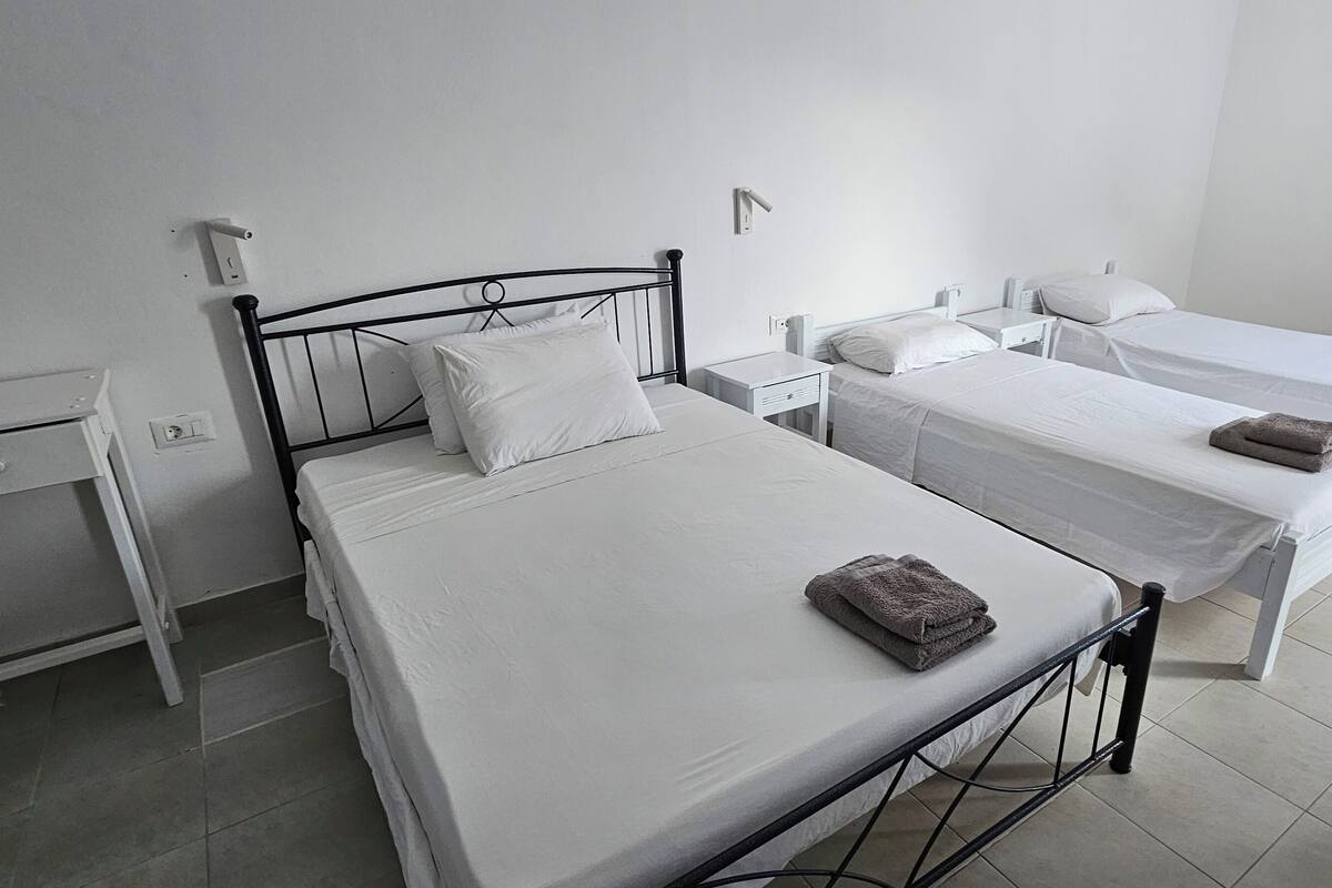 Habitación moderna y limpia con tres camas individuales, cada una con colchón y almohadas blancos a juego. Cada cama tiene una mesita de noche pequeña con una lámpara. Las paredes son blancas y el suelo, de baldosas.
