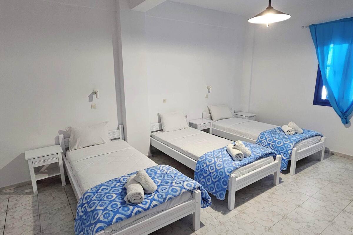 Una habitación con tres camas individuales, cada una con colchón blanco y ropa de cama de diseño azul. Hay mesitas de noche y una ventana con cortinas azules.