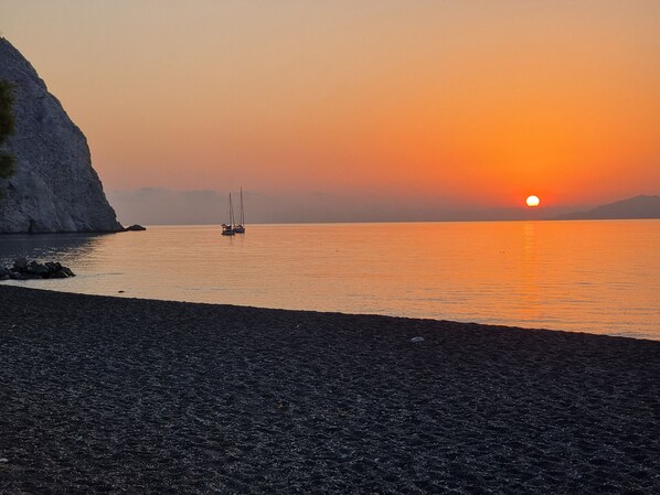 Ein Sonnenuntergang über einem ruhigen Meer mit einer felsigen Küstenlinie und einem Segelboot in der Ferne.