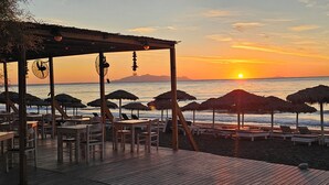 Un restaurant situé en bord de mer, avec des tables et des chaises en bois, des parasols en chaume et une terrasse en bois offrant une vue sur l’océan.