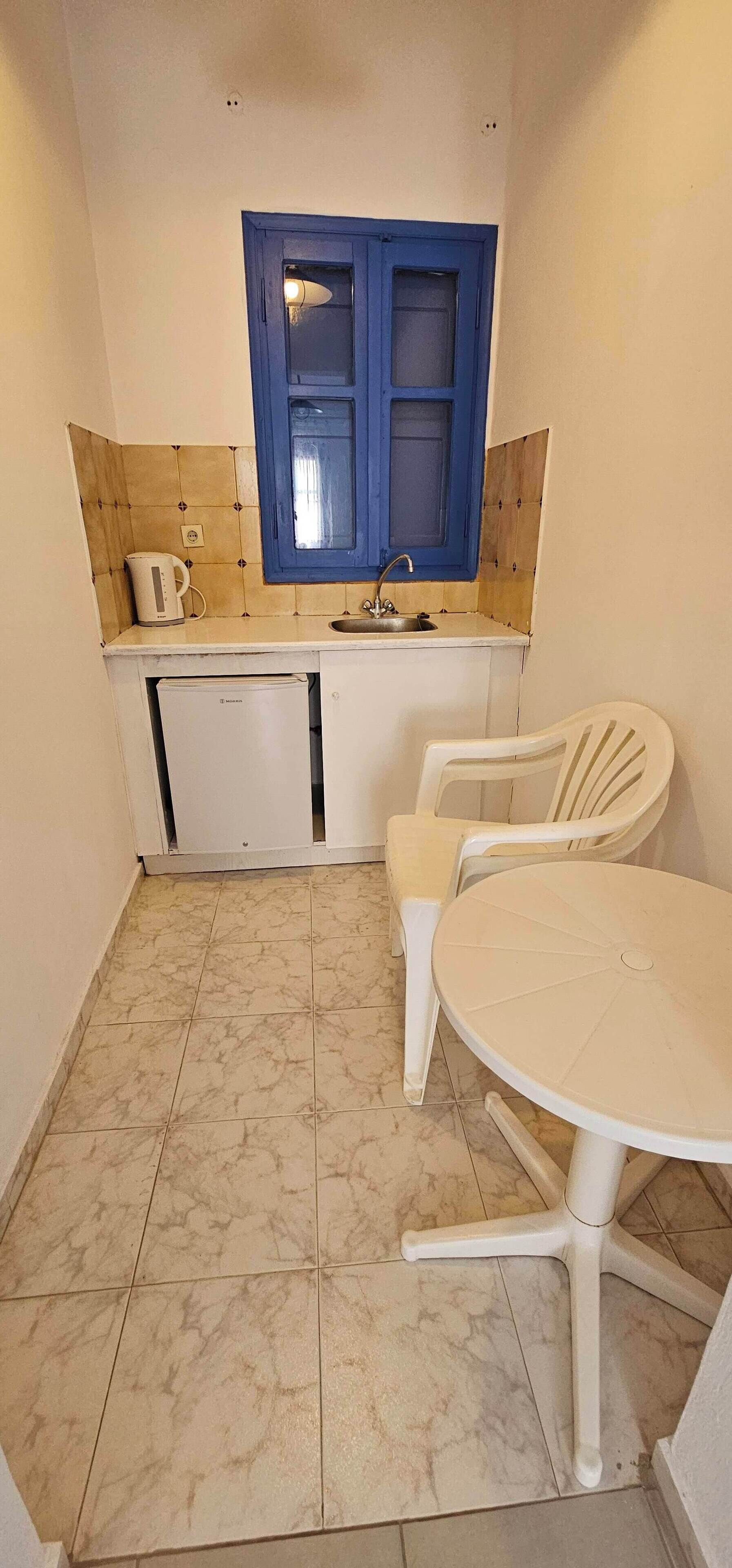 A compact kitchen with a blue-framed window, a sink, a white kettle, and a small refrigerator. There is a white plastic chair and a round table with a white chair.