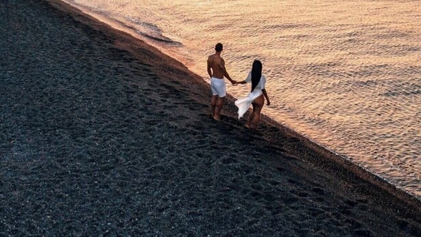 Une scène de plage où un couple se tient la main près du bord de l’eau, avec des bateaux à l’horizon et une falaise rocheuse sur la gauche.