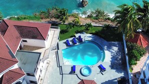 An aerial view of a resort with a swimming pool surrounded by lounge chairs, a rocky outcrop in the sea, and palm trees.