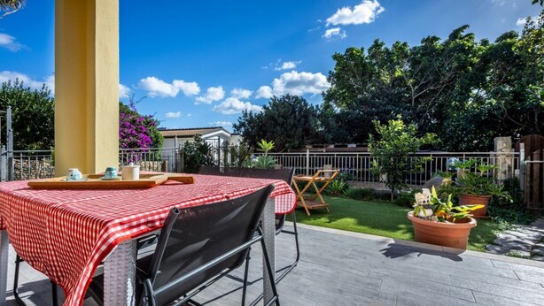 Um pátio ao ar livre com uma mesa posta para dois, um pilar amarelo e um jardim com plantas em vasos.