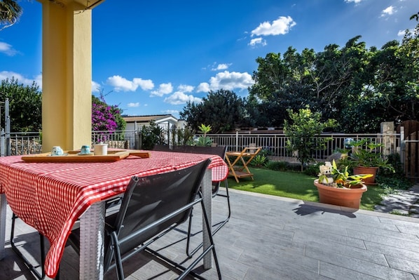 An outdoor patio with a table set for two, a yellow column, and a garden with potted plants.