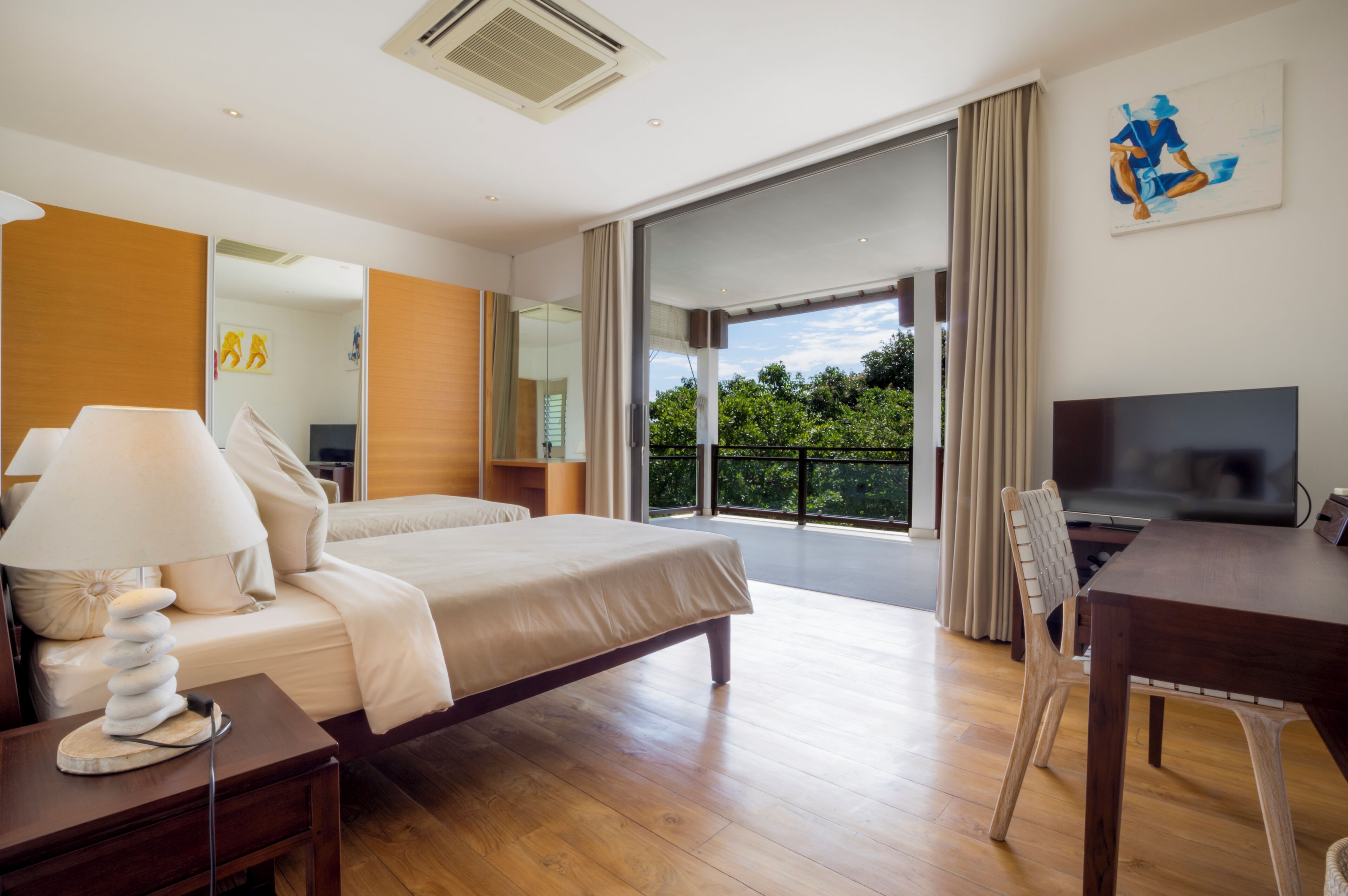 A modern hotel room with a large bed, a flat-screen TV, a dining table, and a balcony with a view of greenery.