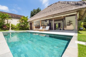 Ein Pool mit Liegestühlen, eine strohgedeckte Struktur und üppiges Grün.