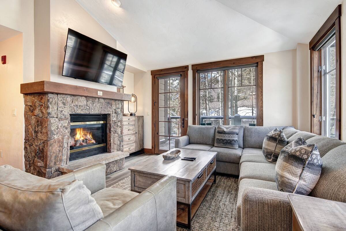 A living room with a stone fireplace, a flat-screen TV mounted above it, a sectional sofa, and a coffee table.