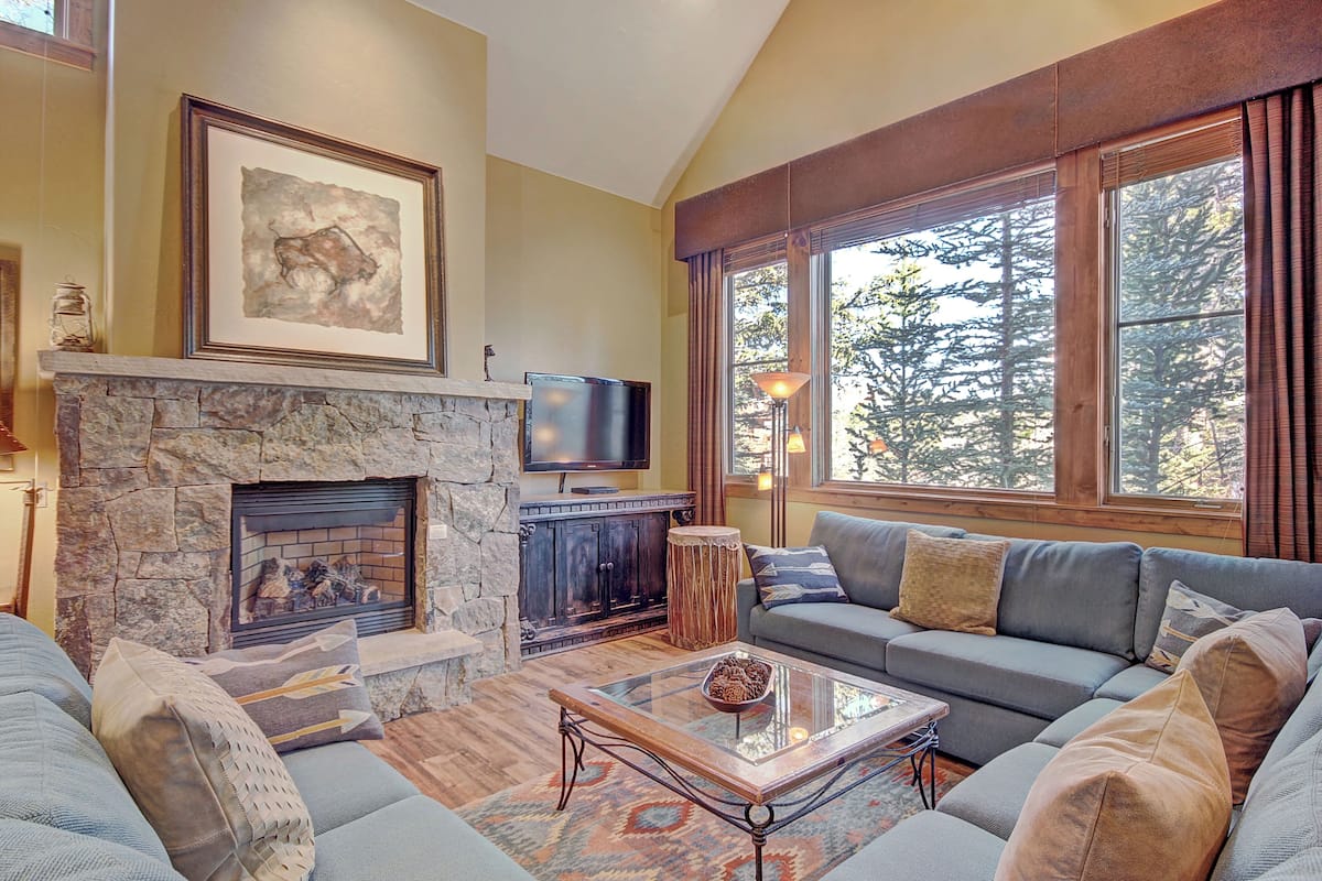 A cozy living room with a stone fireplace, a large window with a forest view, a comfortable sofa, and a flat-screen TV.