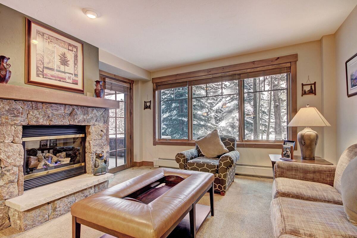 A cozy living room with a stone fireplace, large windows with a snowy view, a comfortable armchair, and a lamp on a side table.