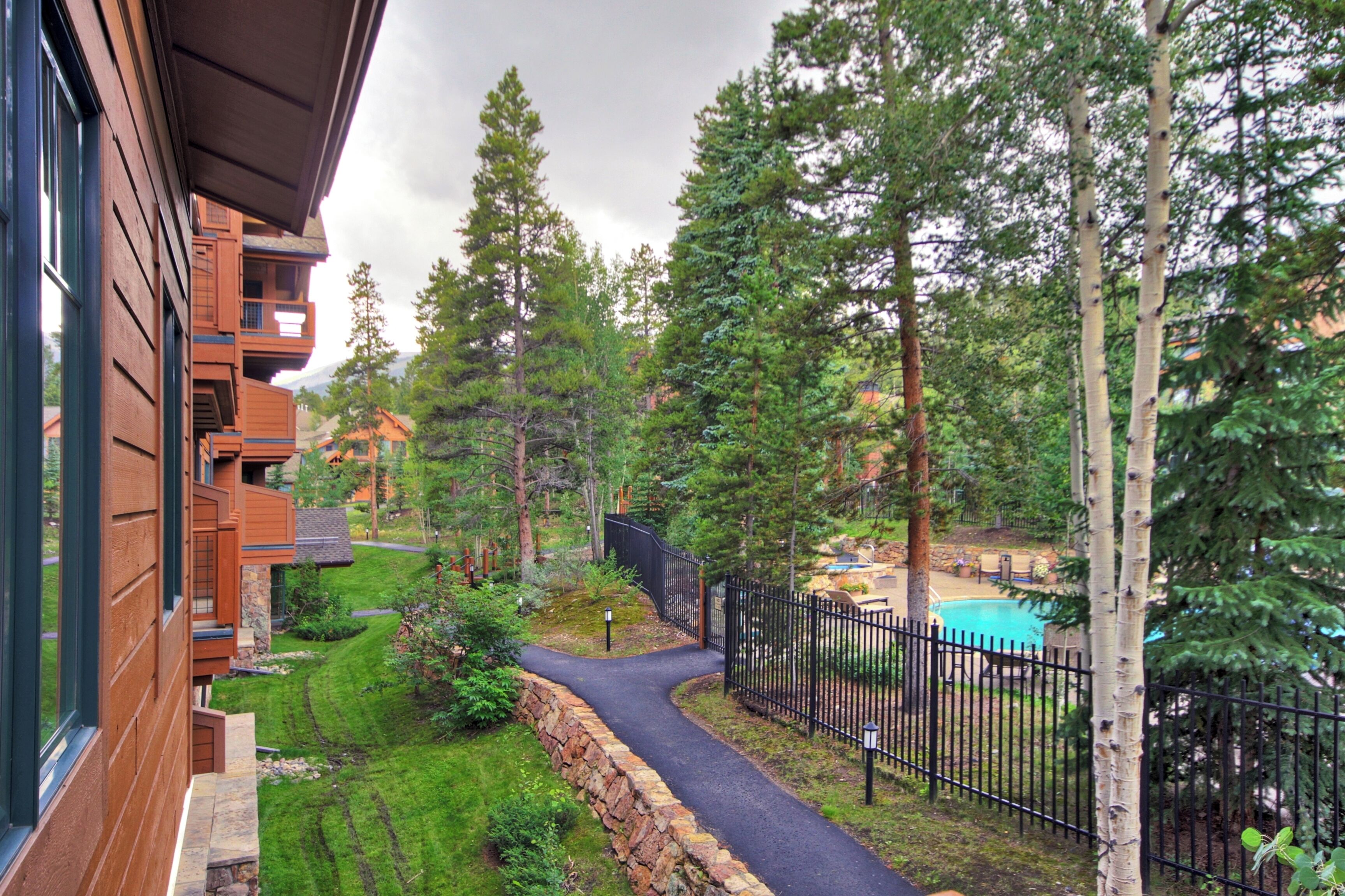 A residential building with a stone pathway leading to a swimming pool, surrounded by trees and a well-maintained lawn.
