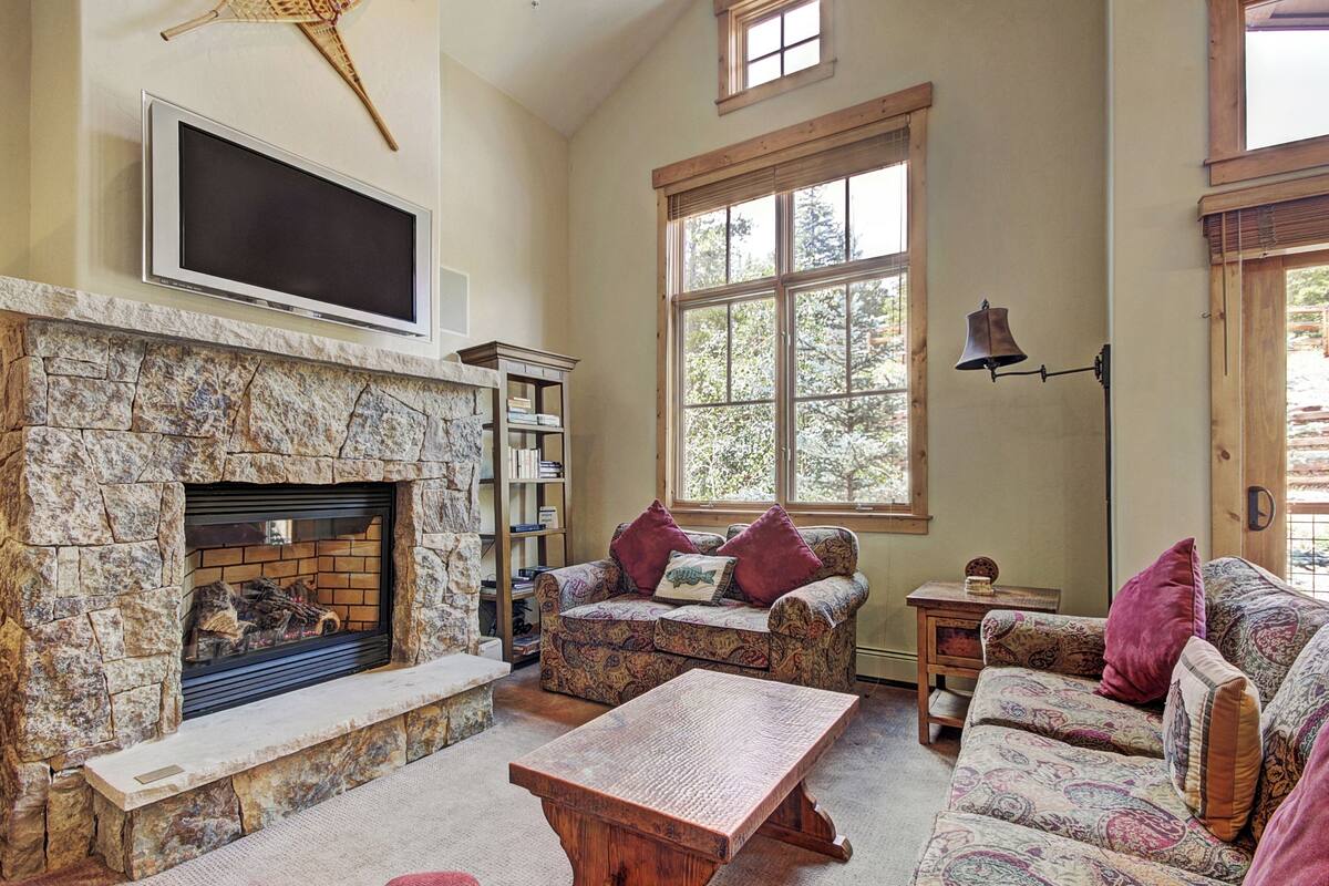 A living room with a stone fireplace, patterned sofas, a flat-screen TV, and large windows.