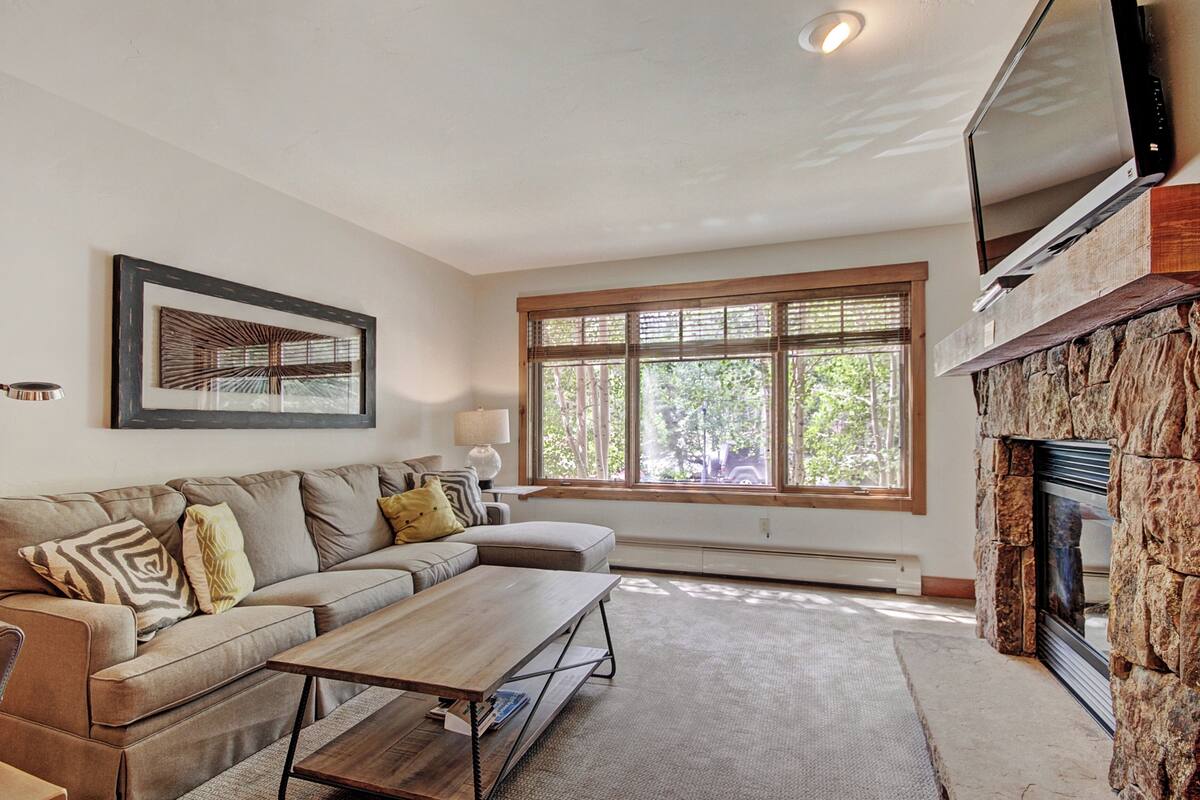 A living room with a sectional sofa, a coffee table, a fireplace, a television, and a large window with blinds.