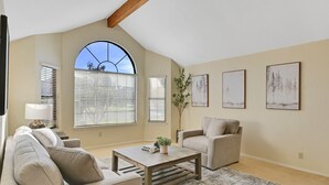 Una sala de estar con techo alto, ventanas grandes con persianas, un sofá, una mesa de centro y cuadros en las paredes.