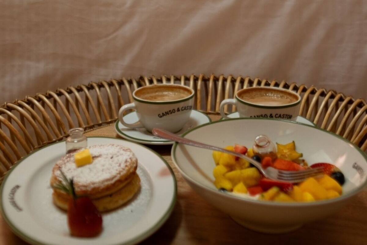 Une table de petit-déjeuner avec deux tasses étiquetées « GANSO & CASTOR », une assiette avec une pâtisserie et un petit fruit rouge, et un bol de fruits mélangés. En arrière-plan, deux personnes sont assises sur un lit, une serviette blanche sur la tête.