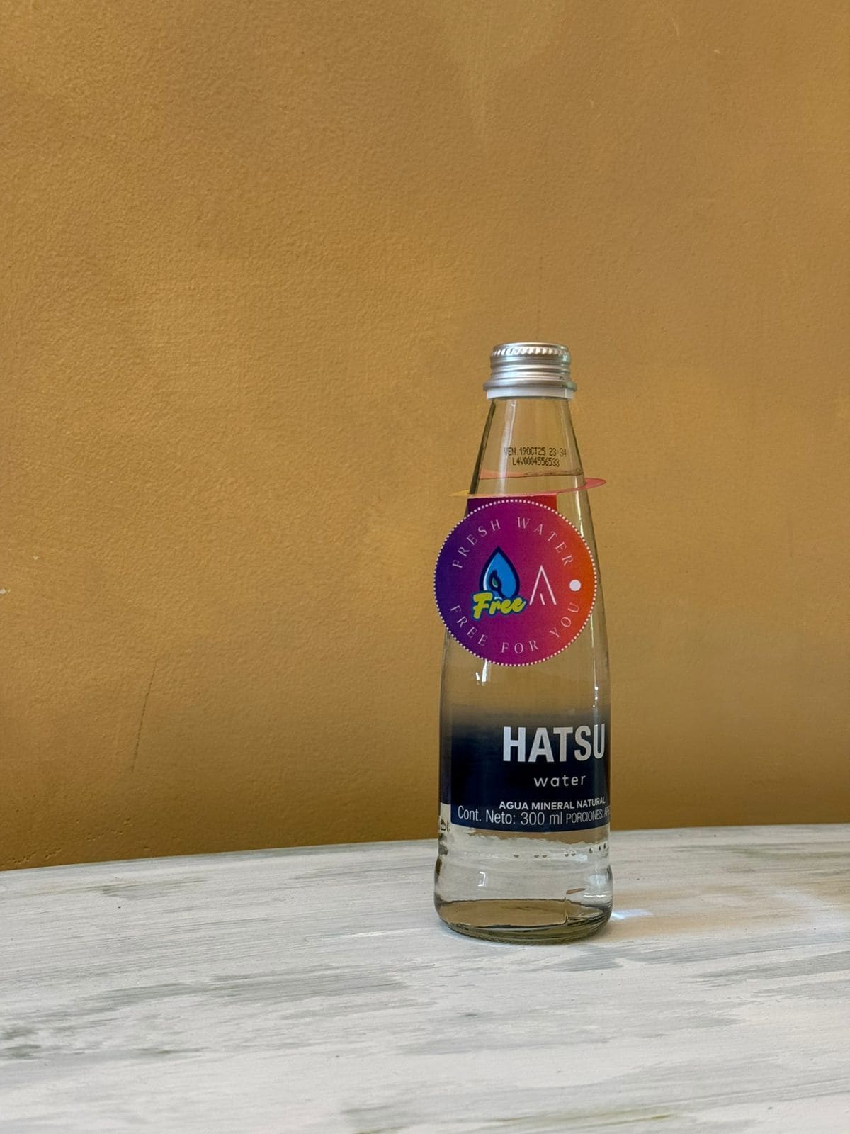 A clear glass bottle of Hatsu water with a purple label, placed on a marble surface against a plain background.