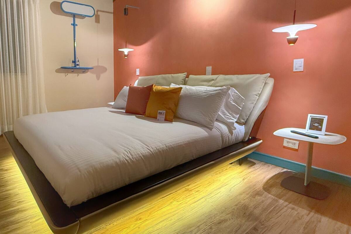 Un lit bien fait, avec du linge de lit blanc, une table de chevet avec une lampe et un luminaire fixé au mur.