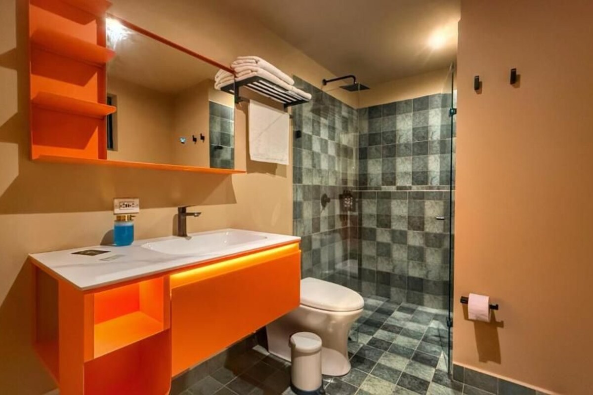 Un baño con lavamanos blanco, mueble naranja y ducha con azulejos a cuadros.